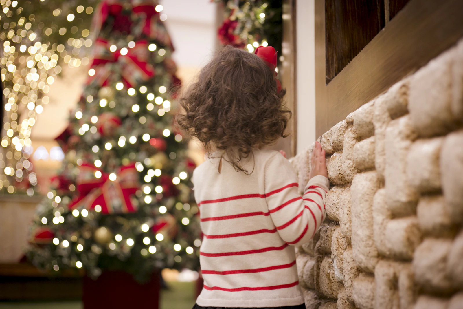 Petite fille de dos contre un mur en pierre, sapin de Noël illuminé derrière