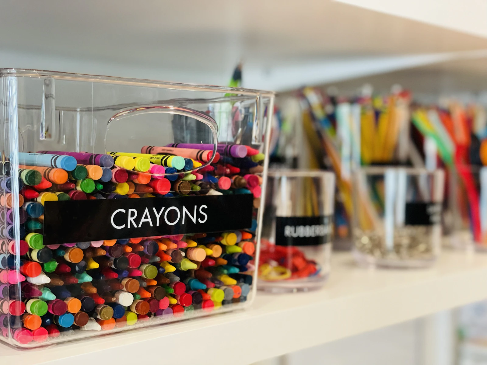 Bac transparent étiqueté rempli de crayons de cire sur une étagère blanche