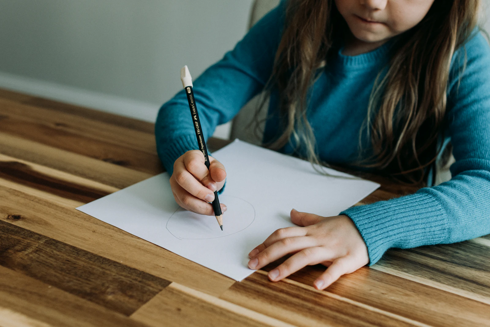 Fillette en pull bleu traçant au crayon sur une feuille blanche posée sur une table